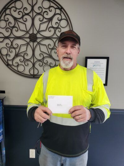 Man in safety vest holding a white envelope; in an office setting, decorative wall art behind him. | Joe's Slinger Service