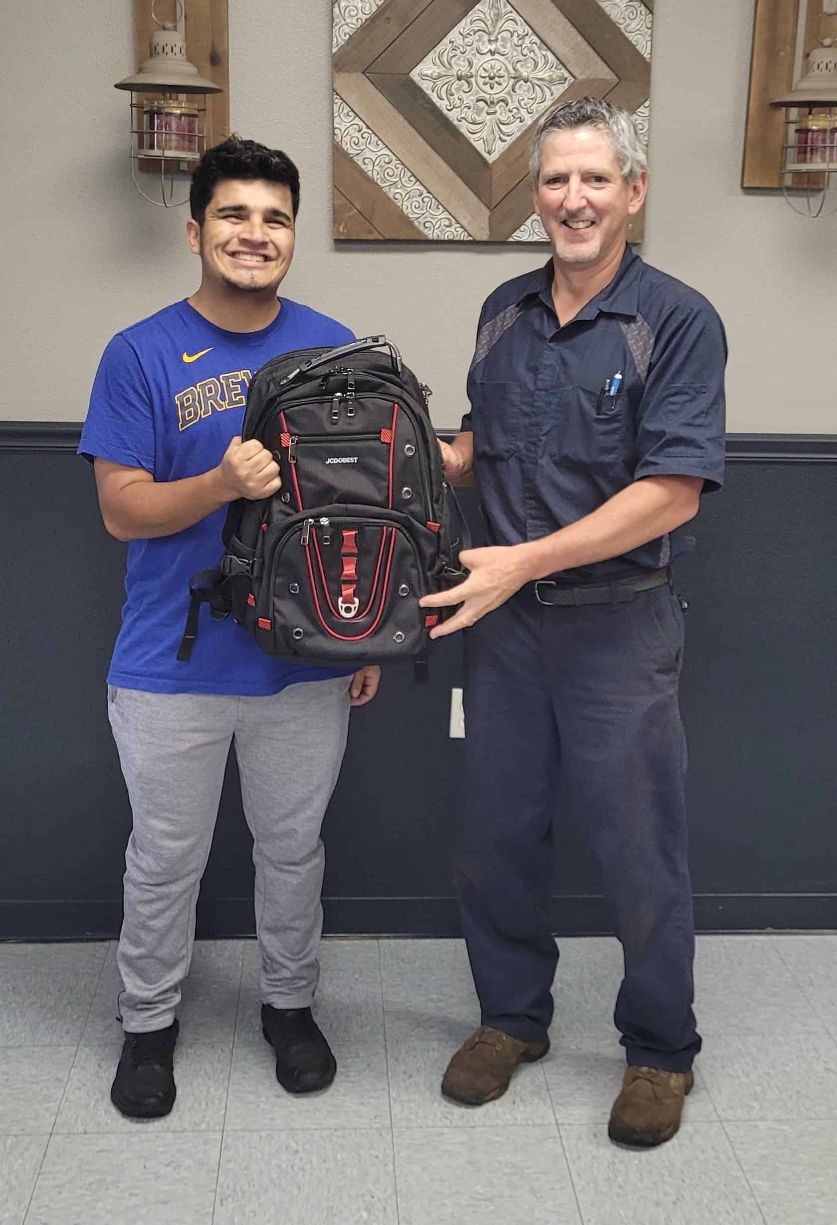 Man holding a backpack smiles, next to another man in a blue shirt smiling. Indoors, against a wall. | Joe's Slinger Service