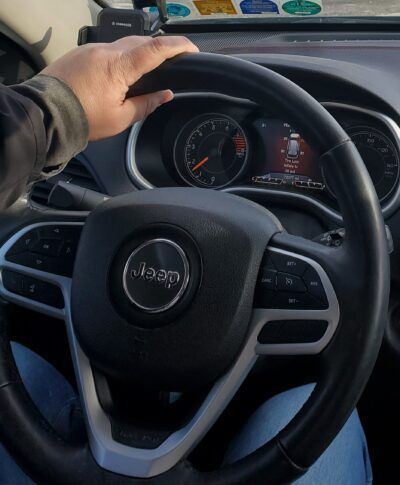 Person's hand on a Jeep steering wheel, inside the car. Dashboard and gauges are visible. | Joe's Slinger Service