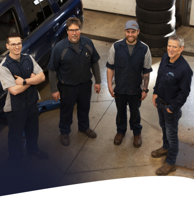 Four mechanics in a garage, wearing work uniforms. The mechanics are standing and looking at the camera. | Joe's Slinger Service