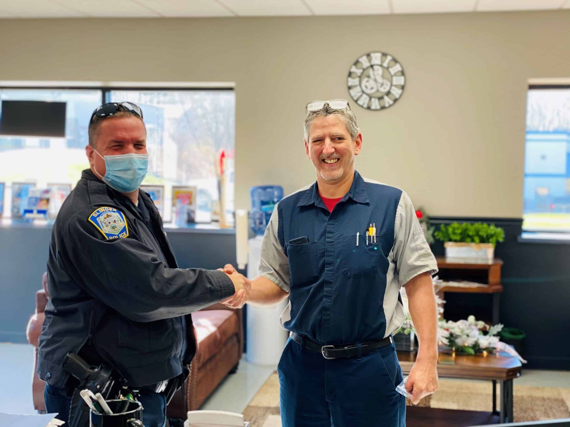 A uniformed officer in a mask shakes hands with a man in mechanic overalls inside an office. | Joe's Slinger Service