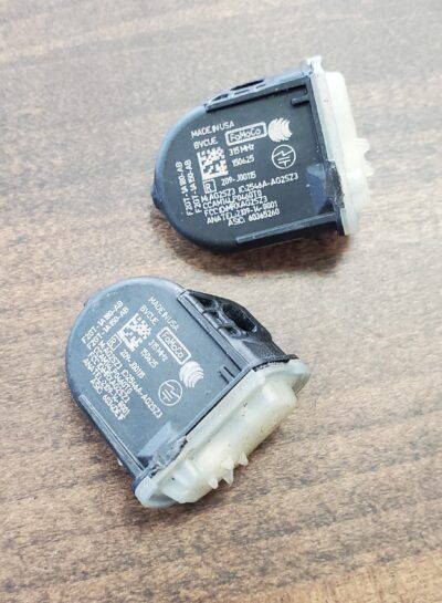 Two black TPMS sensors on a wooden surface, showing labels and white plastic connectors. | Joe's Slinger Service