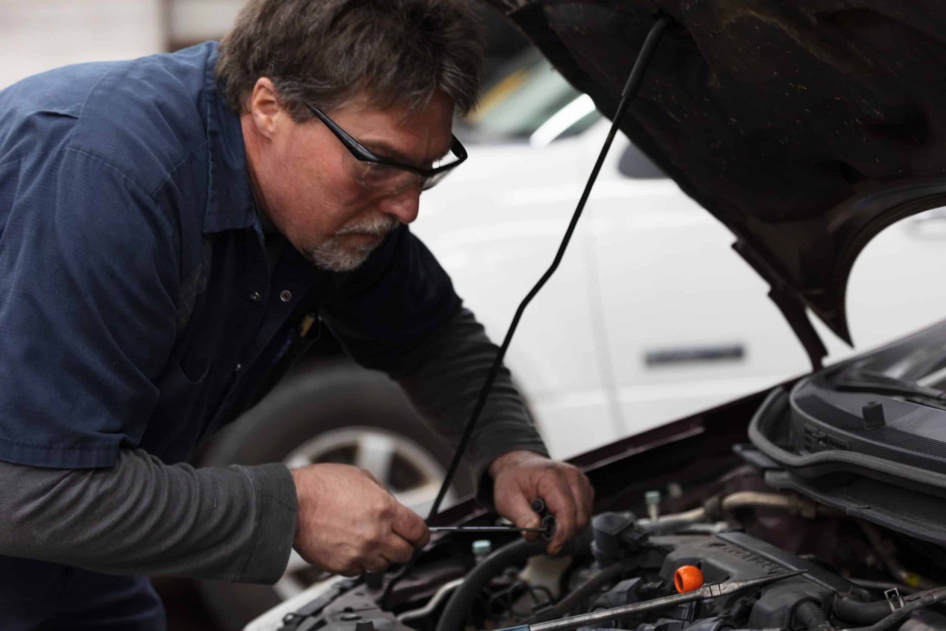 Mechanic working under the hood of a car. Wearing glasses and blue uniform, looking intently. | Joe's Slinger Service