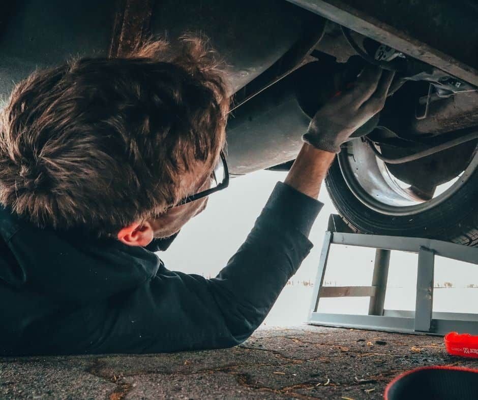 Mechanic working on car undercarriage, wearing gloves, lying on ground near a ramp, inspecting and holding a part. | Joe's Slinger Service
