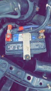 Car battery in an engine bay with connected terminals and a hold-down bracket. | Joe's Slinger Service
