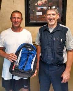 Two men holding a blue and black backpack, smiling indoors near a framed artwork. | Joe's Slinger Service