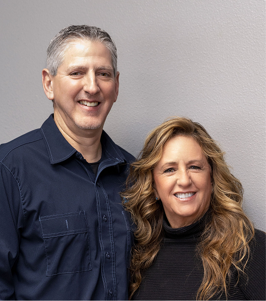 Man and woman smiling, posed in front of decorative wall art. They are indoors. | Joe's Slinger Service