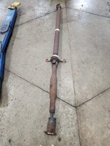Rusty car driveshaft on a concrete floor next to a blue lift. | Joe's Slinger Service