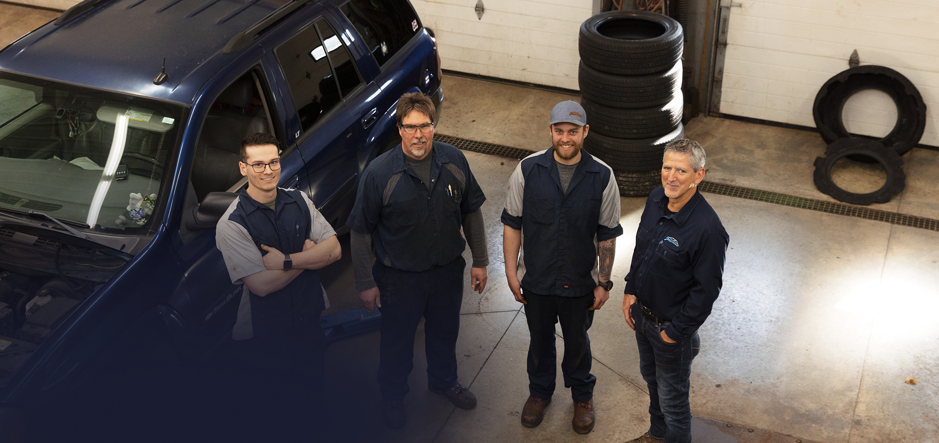 Four men in a garage beside a blue car, smiling. Tires stacked in the background. | Joe's Slinger Service