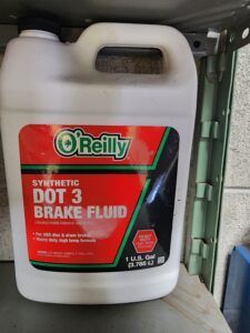 O'Reilly DOT 3 synthetic brake fluid container. Red and green label, white jug, on a metal shelf. | Joe's Slinger Service