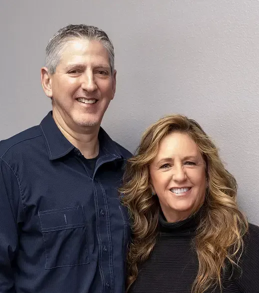 Joe, owner, and his wife smiling, standing in front of a gray wall | Joe's Slinger Service, LLC