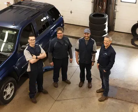 Four auto mechanics standing in a garage beside a blue SUV; one has arms crossed.