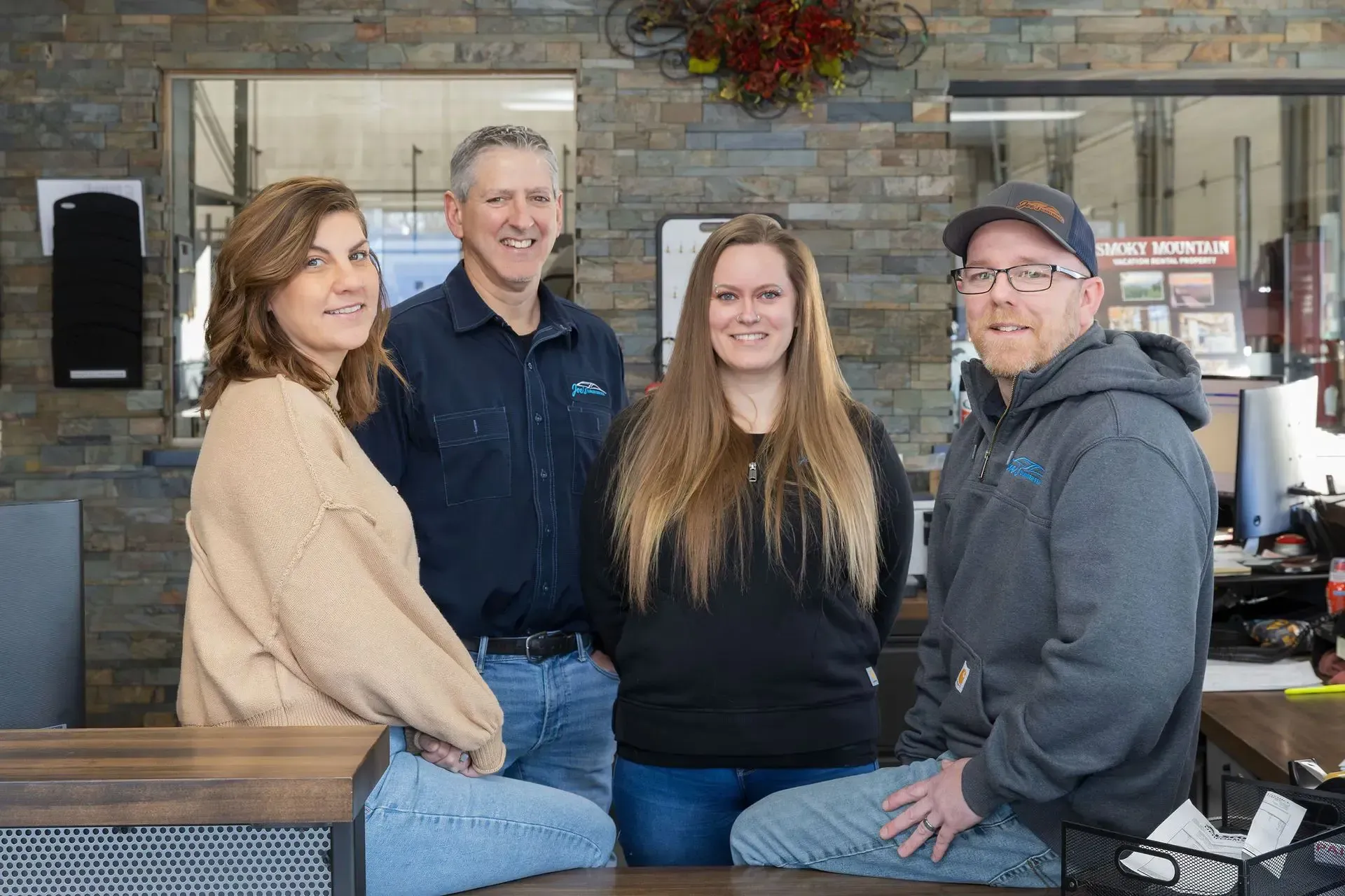 Four people smiling behind a counter inside a business. Brick wall, sunlight, and a decorative wreath.