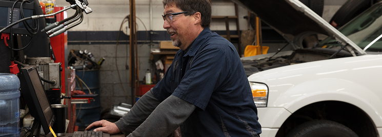 Mechanic at computer in auto repair shop. White car with open hood, tools, and equipment visible. | Joe's Slinger Service