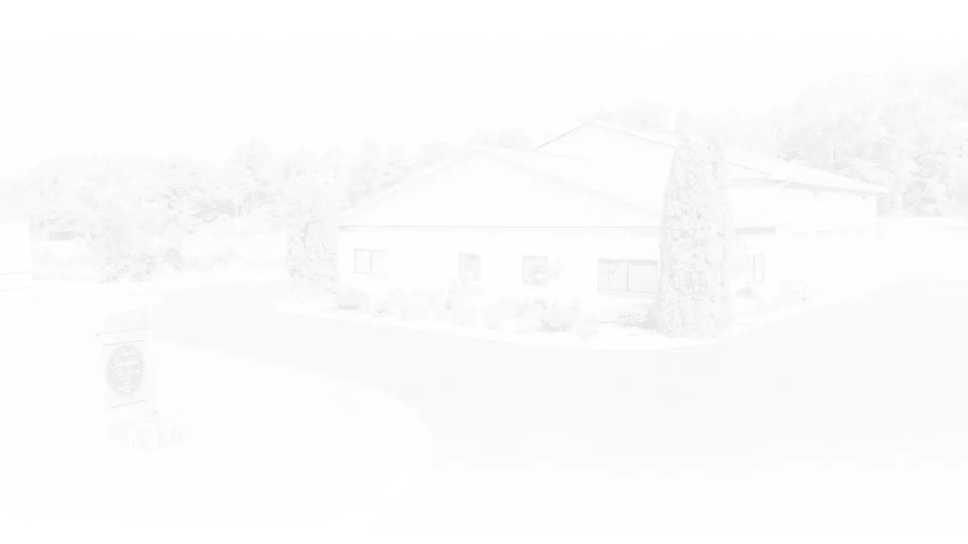 White building with trees, driveway, and a partially visible sign on a bright day.
