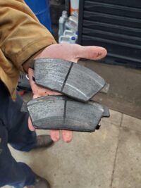 Person holding two worn brake pads. Indoors with a toolbox visible. | Joe's Slinger Service