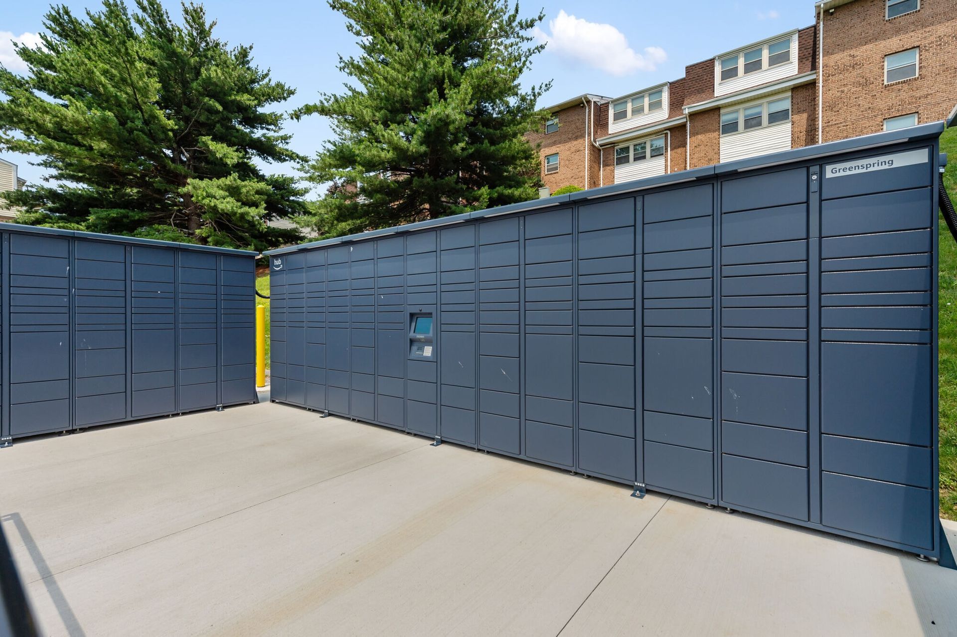 Row of blue storage lockers on a concrete pad with trees and a brick building in the background.