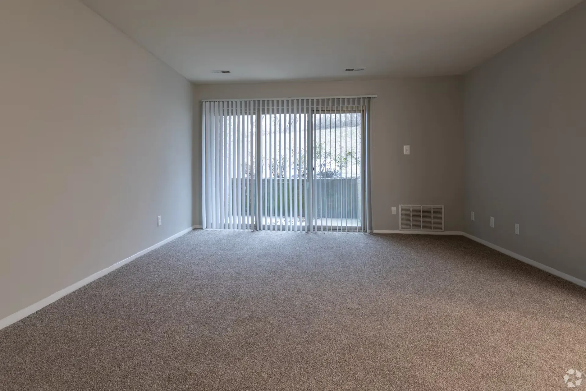 Empty living room with beige walls, carpet, and a sliding glass door with vertical blinds.