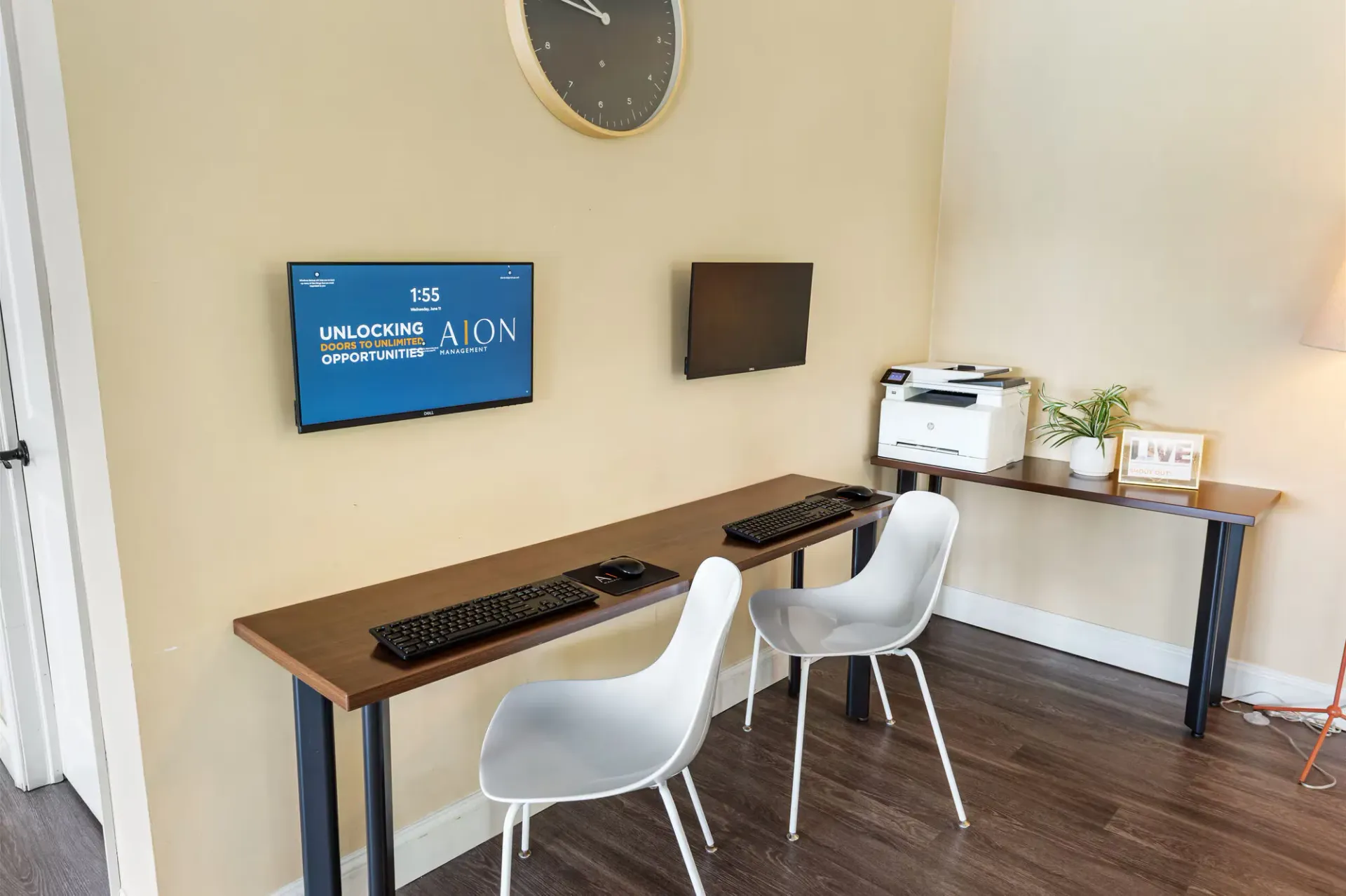 Leasing center workstation with two wall-mounted monitors, a printer, and white chairs in a beige room.