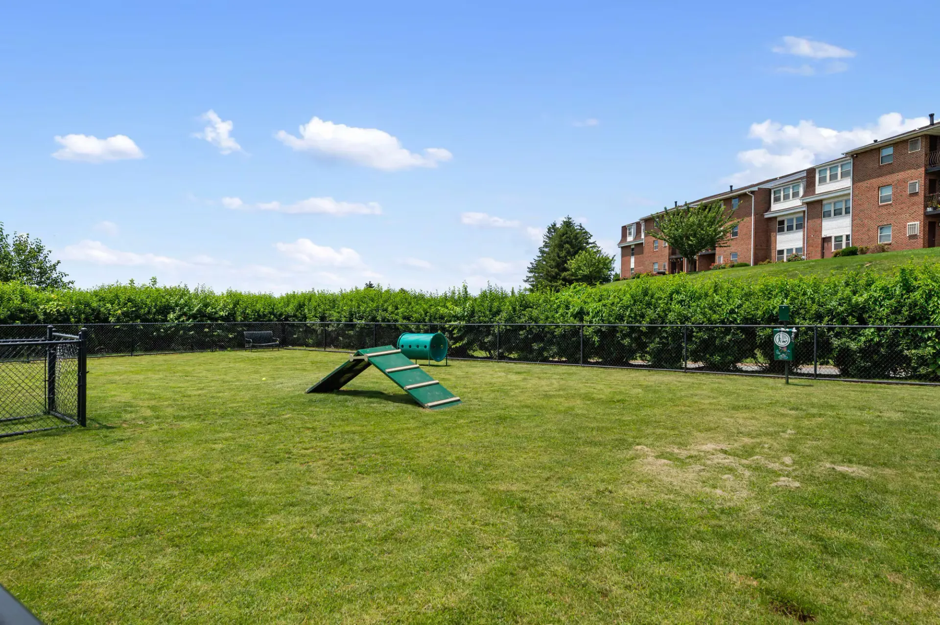 Open grassy community yard with a small green play ramp and tunnel, fenced, beside brick apartment buildings.