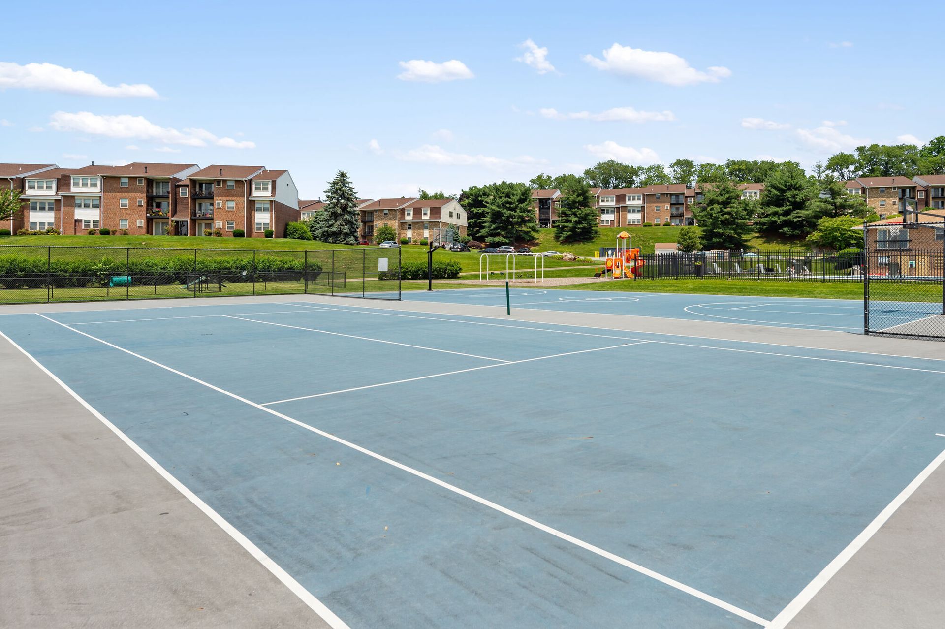 Blue outdoor multi-use court with white lines, fenced, and apartment buildings in the background.