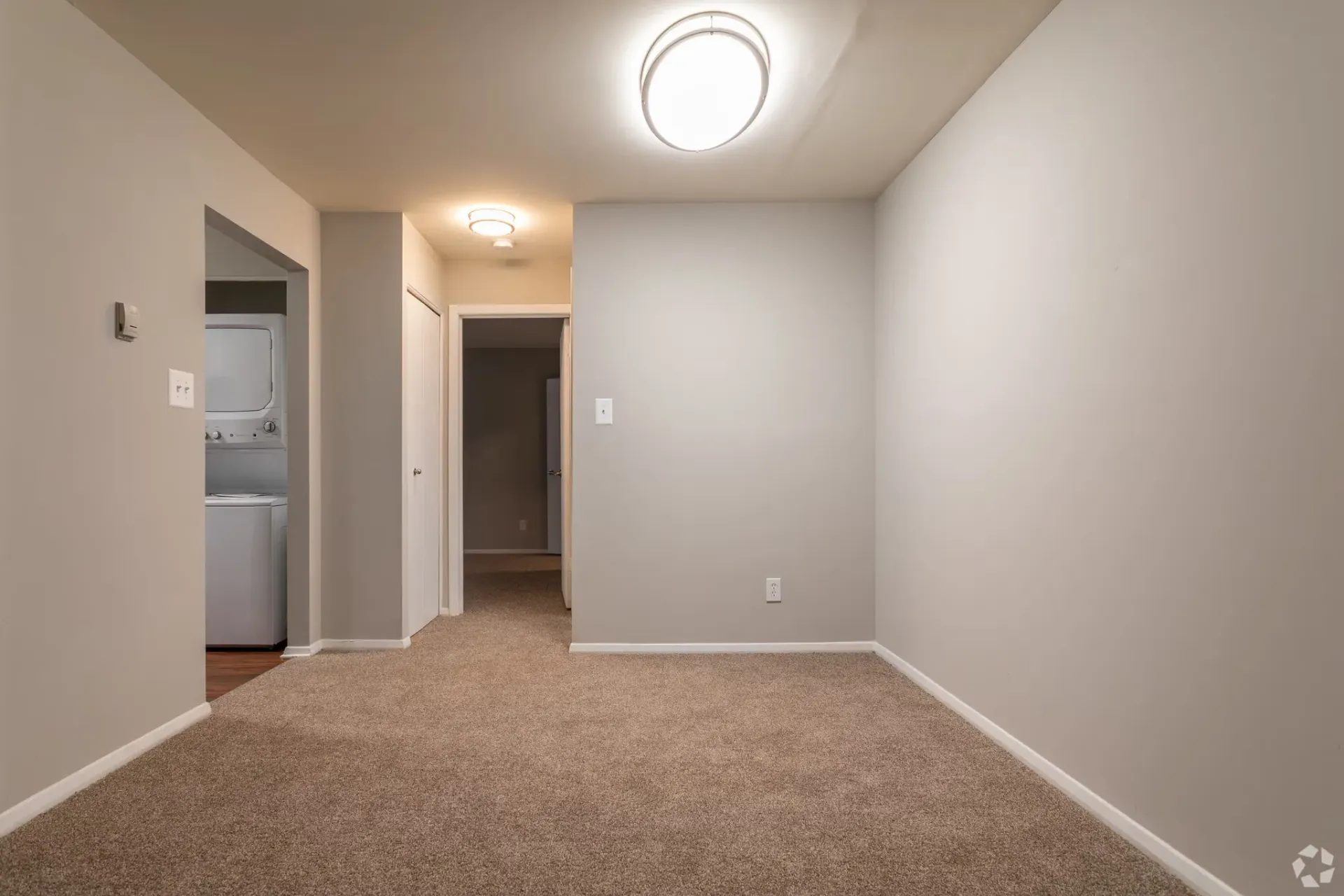 Carpeted apartment living area with a stacked washer/dryer in a left alcove.
