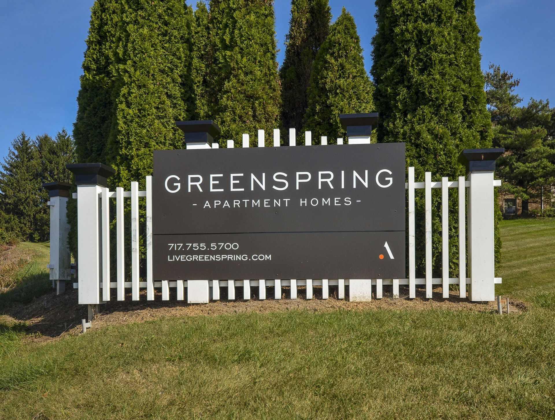 Entrance sign for Greenspring Apartment Homes with white fence and tall evergreen trees.