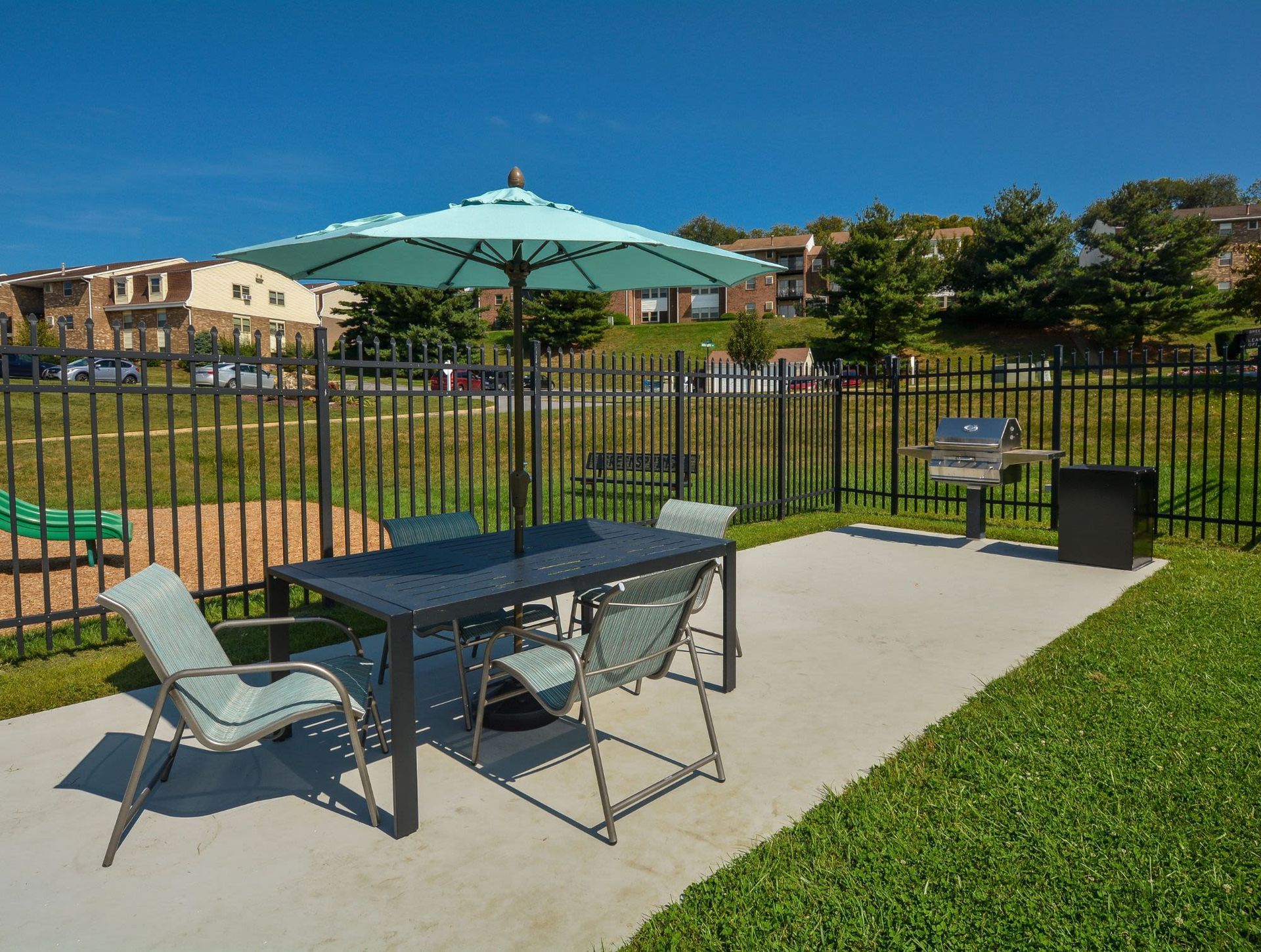 Outdoor amenity area with a table, chairs, umbrella, and a grill behind a black metal fence.