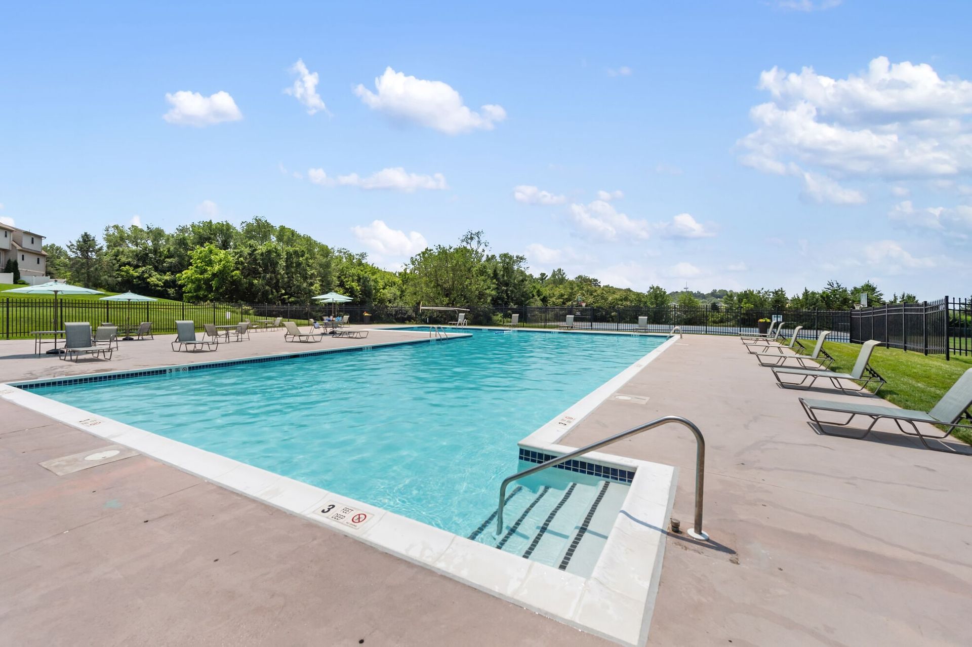 Outdoor community pool with lounge chairs, umbrellas, and a fence.