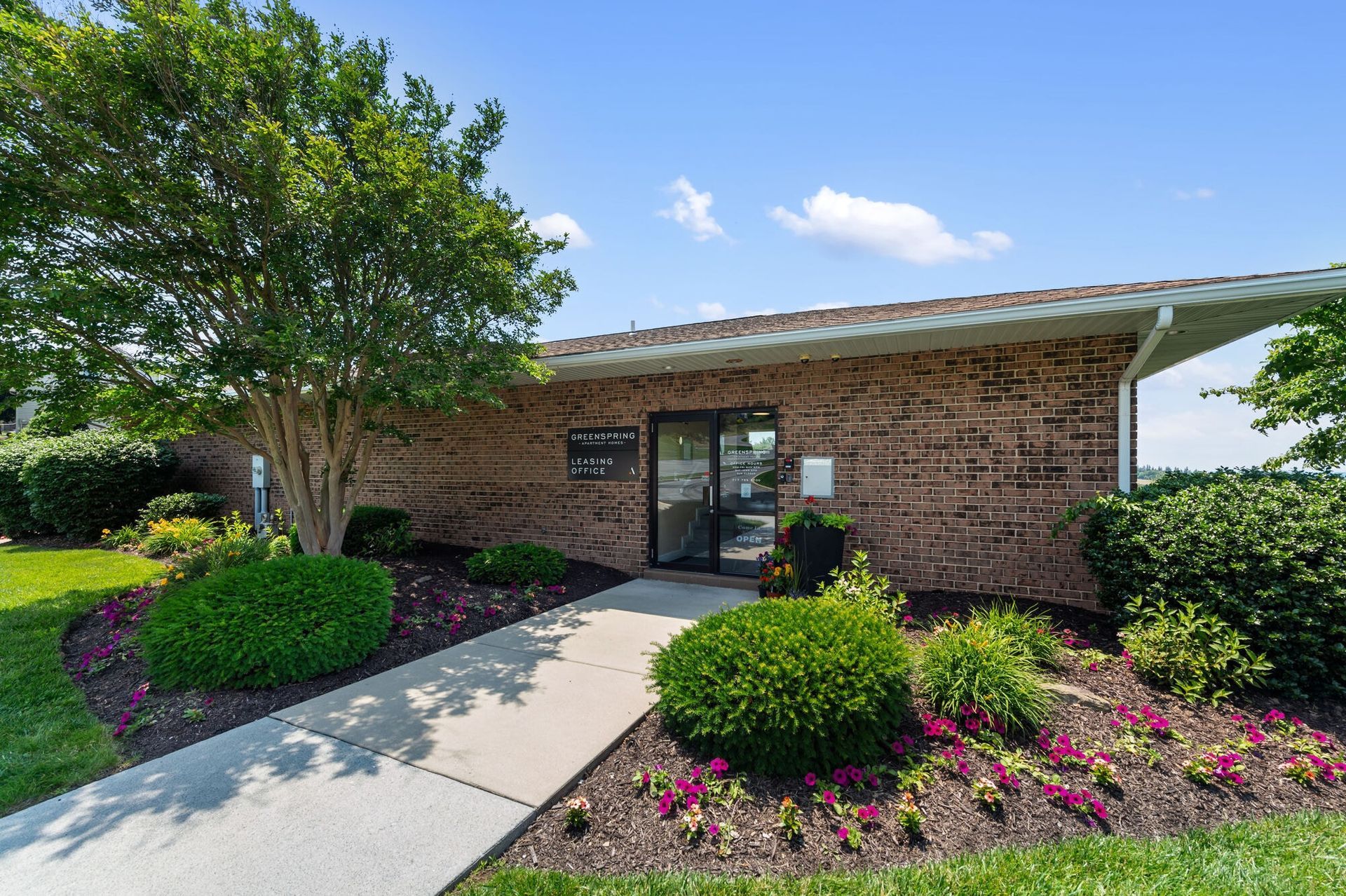 Brick leasing office entrance with glass door, flanked by shrubs and a tree.