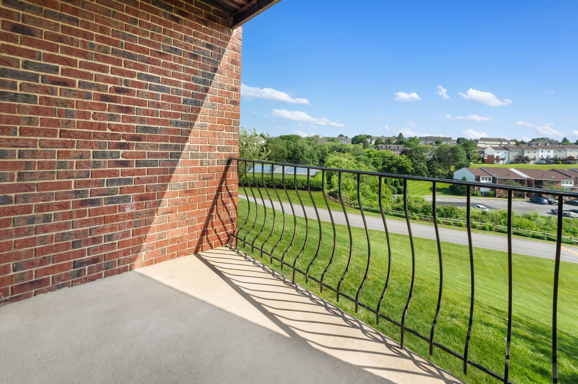Balcony with brick wall and curved black railing, overlooking a green lawn and distant houses.