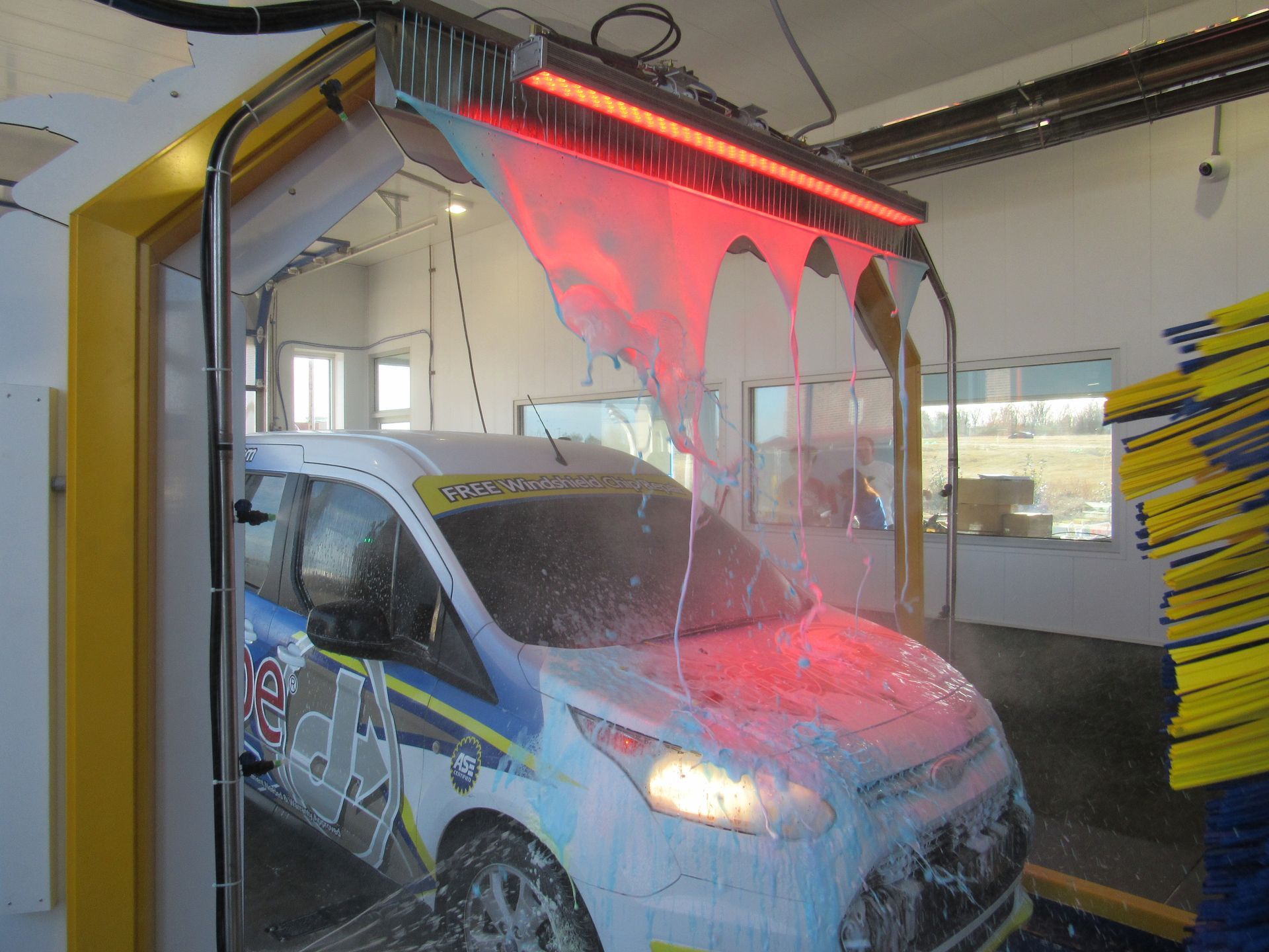 A white van with racing decals being washed in a car wash bay, with red illuminated water curtains.