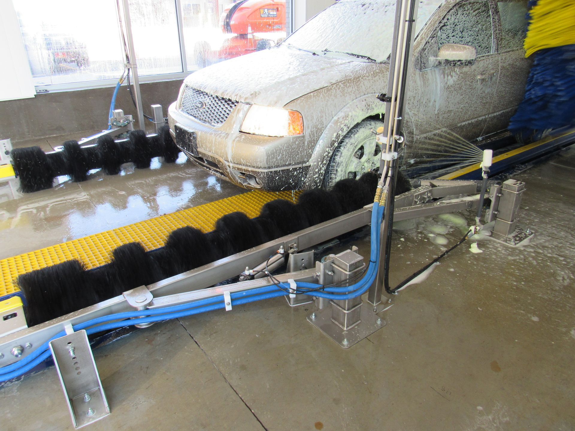 Car being washed at a car wash. A vehicle is on a conveyor belt and covered in soap with brushes cleaning its sides.