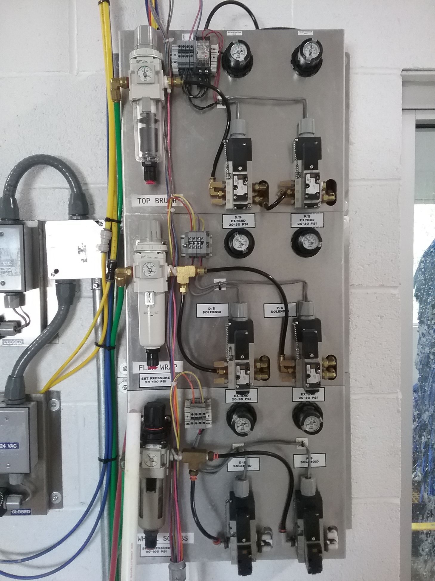 A silver pneumatic control panel mounted on a wall with gauges, valves, and filters, various colored wires, and tubing.