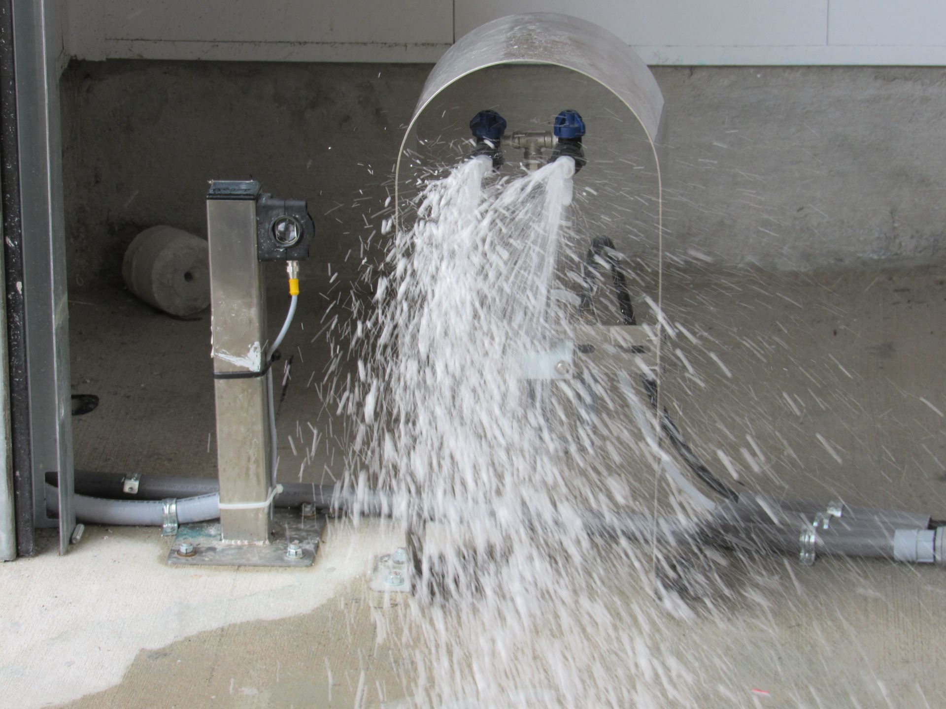 A stainless steel emergency eye wash station sprays water forcefully onto a concrete floor.