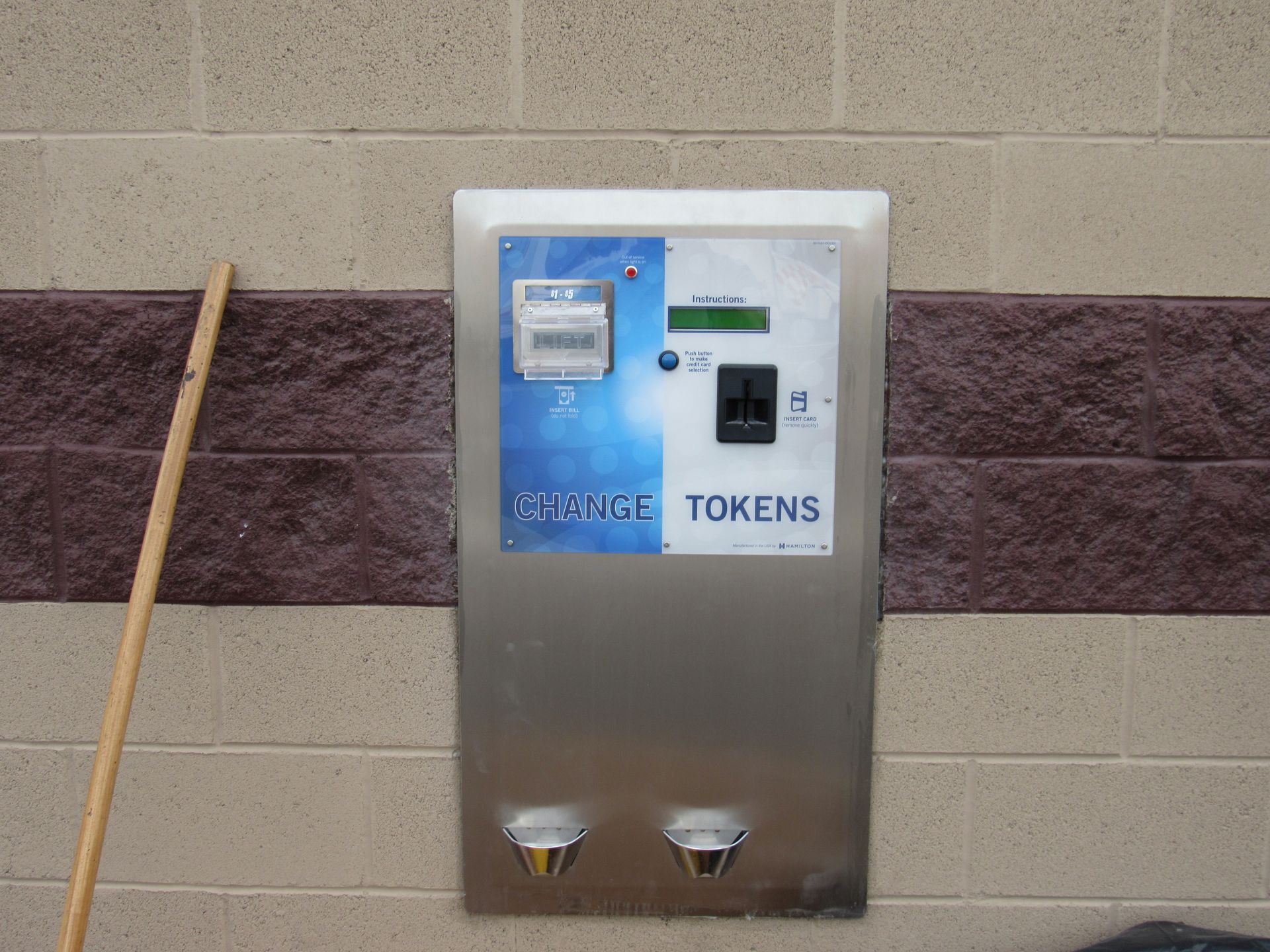 Metal token machine on a brick wall, with blue and white panel and two coin dispensers.