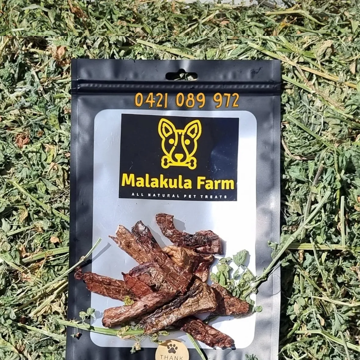 Black pouch of Malakula Farm pet treats, featuring the logo and phone number, with jerky on a grassy background.