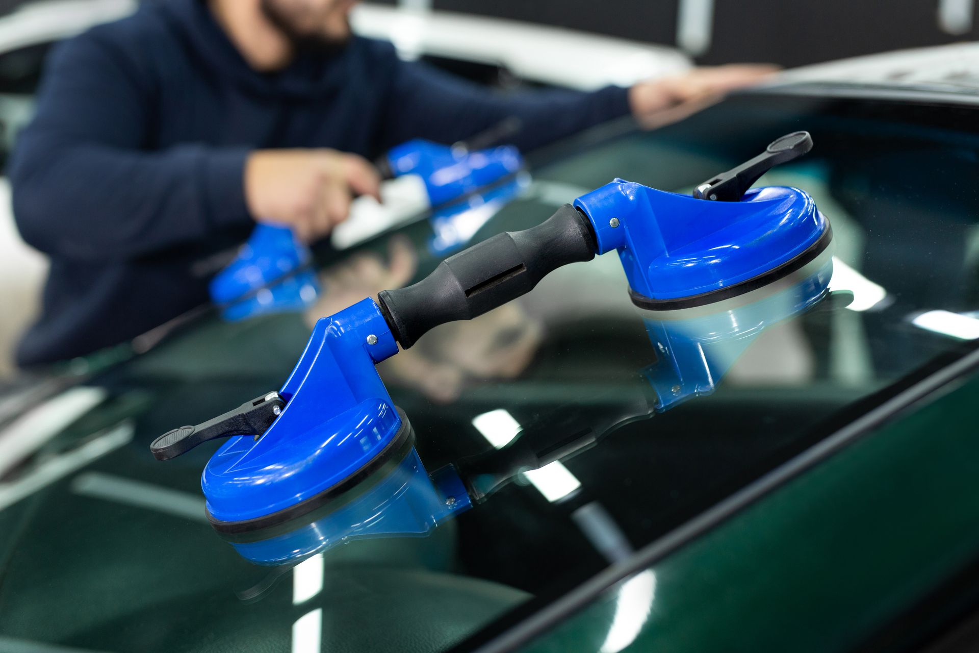 Person using blue suction cups to handle a car windshield in a repair shop.