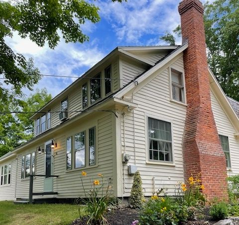 Beige two-story house with a red brick chimney and windows, set against a blue sky with trees and flowers.
