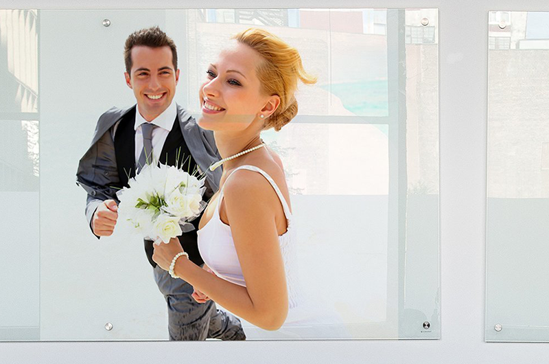 Les mariés sourient en portant un bouquet. Le marié est en costume, la mariée en robe blanche. Le décor est lumineux et ensoleillé.