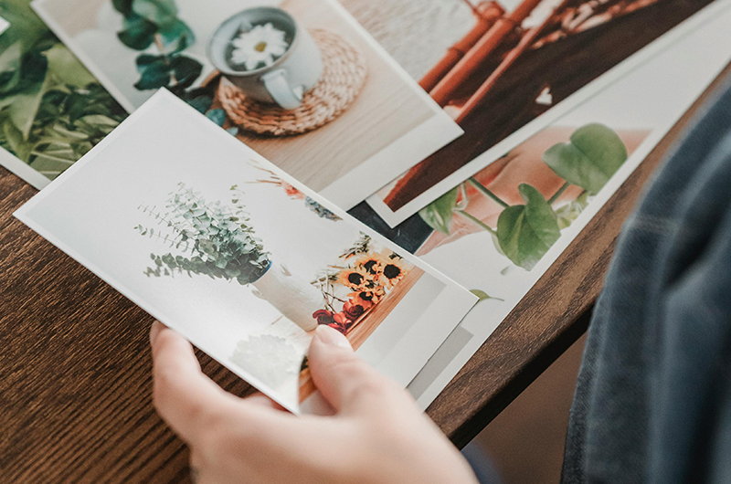 Une main tenant une photo imprimée, entourée d'autres photos sur une table en bois, représentant des plantes et des objets.