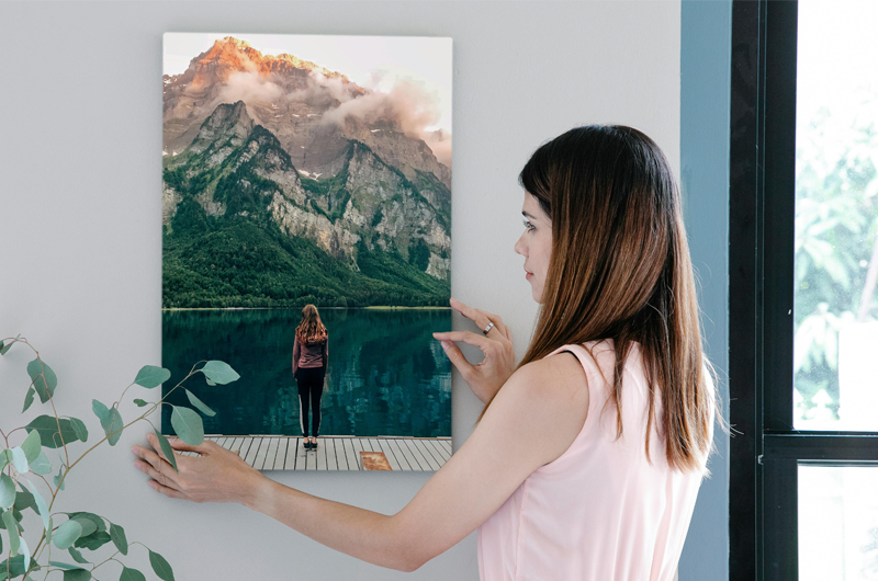 Une femme accroche une toile représentant une personne contemplant un lac et des montagnes.