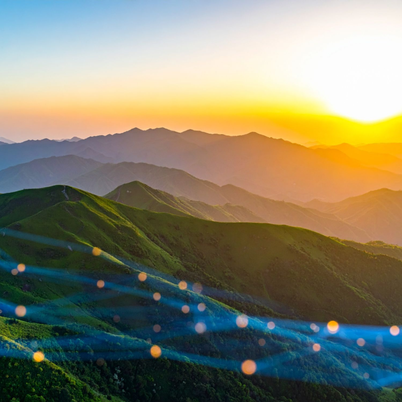 Mountains at sunset, green rolling hills, layers of peaks, orange and blue sky.