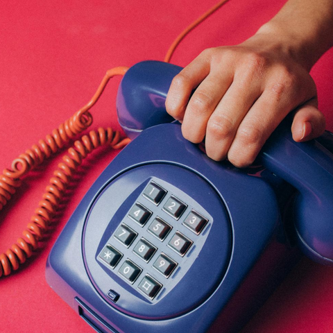 Hand holding a blue push-button telephone on a red background, orange coiled cord visible.