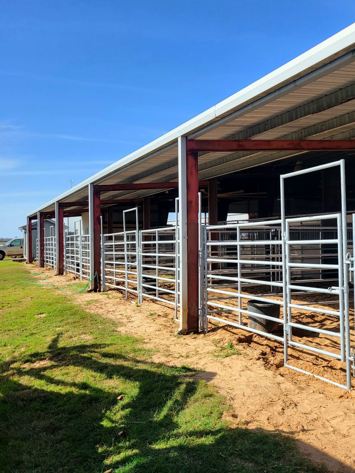 A row of horse pens with a roof and a fence in a field.
