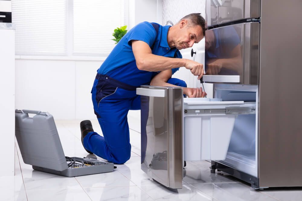 A Man Is Kneeling Down Fixing a Refrigerator in A Kitchen — Care Mechanical Services in Wagga Wagga, NSW