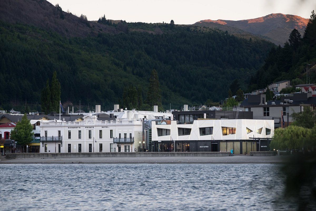 A large white building sits on the shore of a lake with mountains in the background.