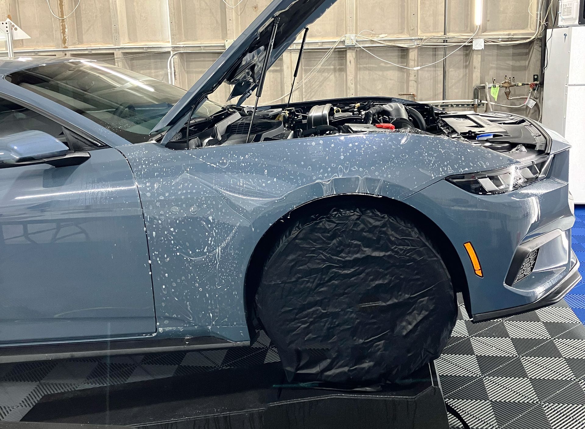 Blue car with hood open and front wheel covered in a repair shop