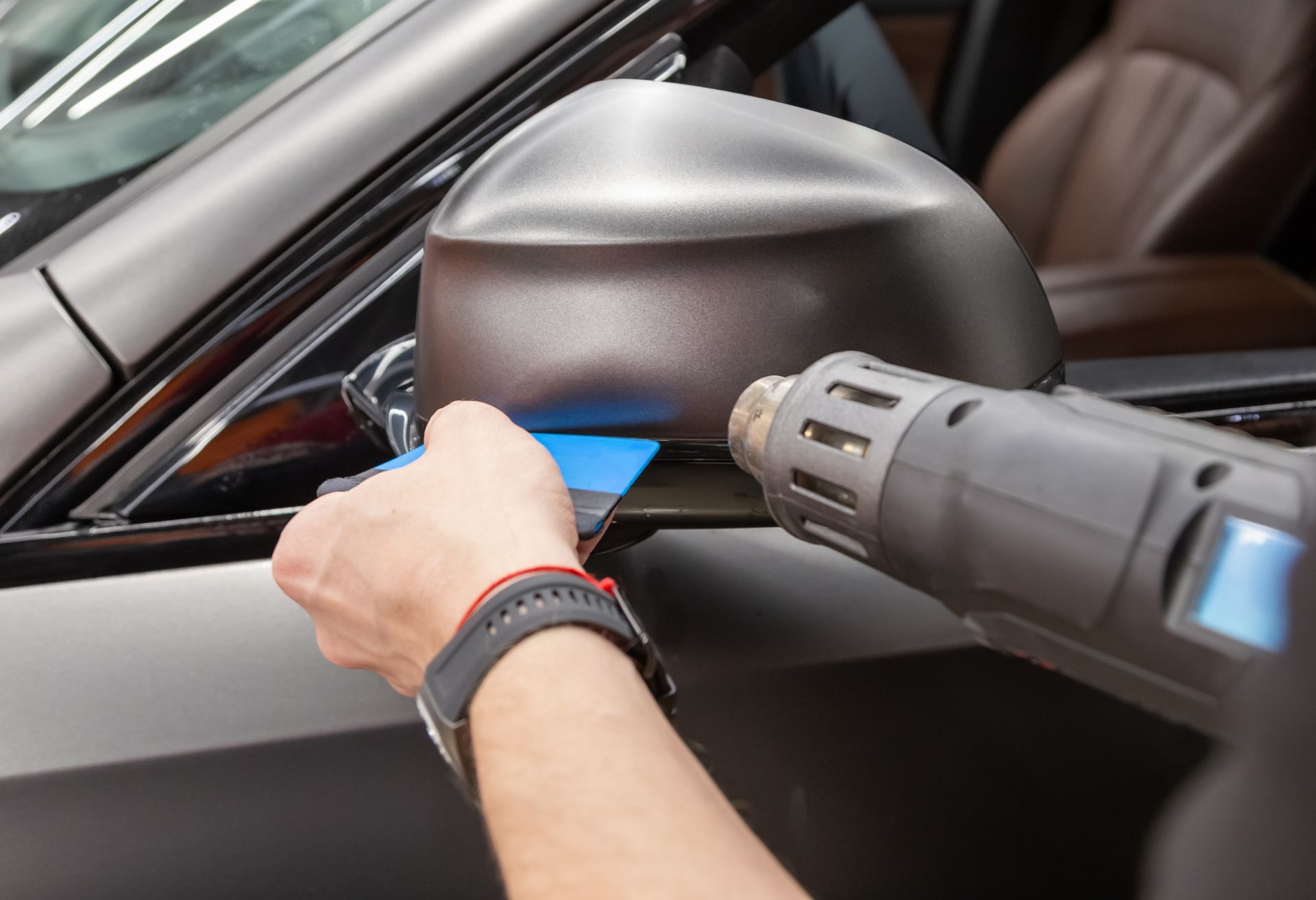 A technician uses a heat gun and a squeegee to apply a matte vinyl wrap to a car’s side mirror.