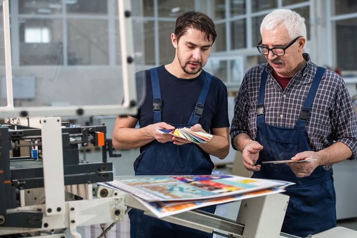 Two men are standing next to each other in a factory looking at a piece of paper.